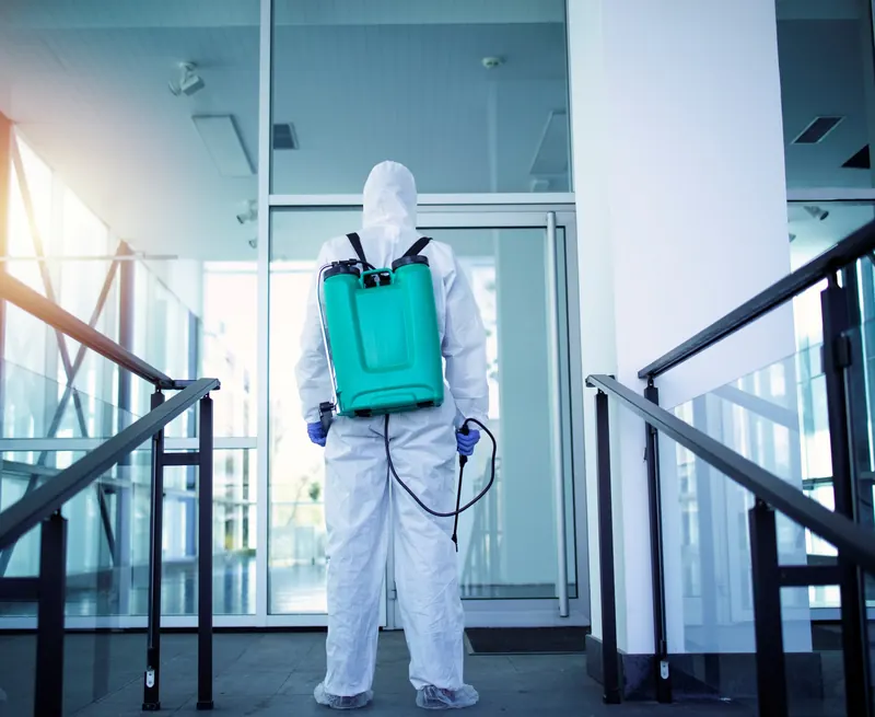 Técnico con traje de protección y mochila pulverizadora en el acceso de un edificio